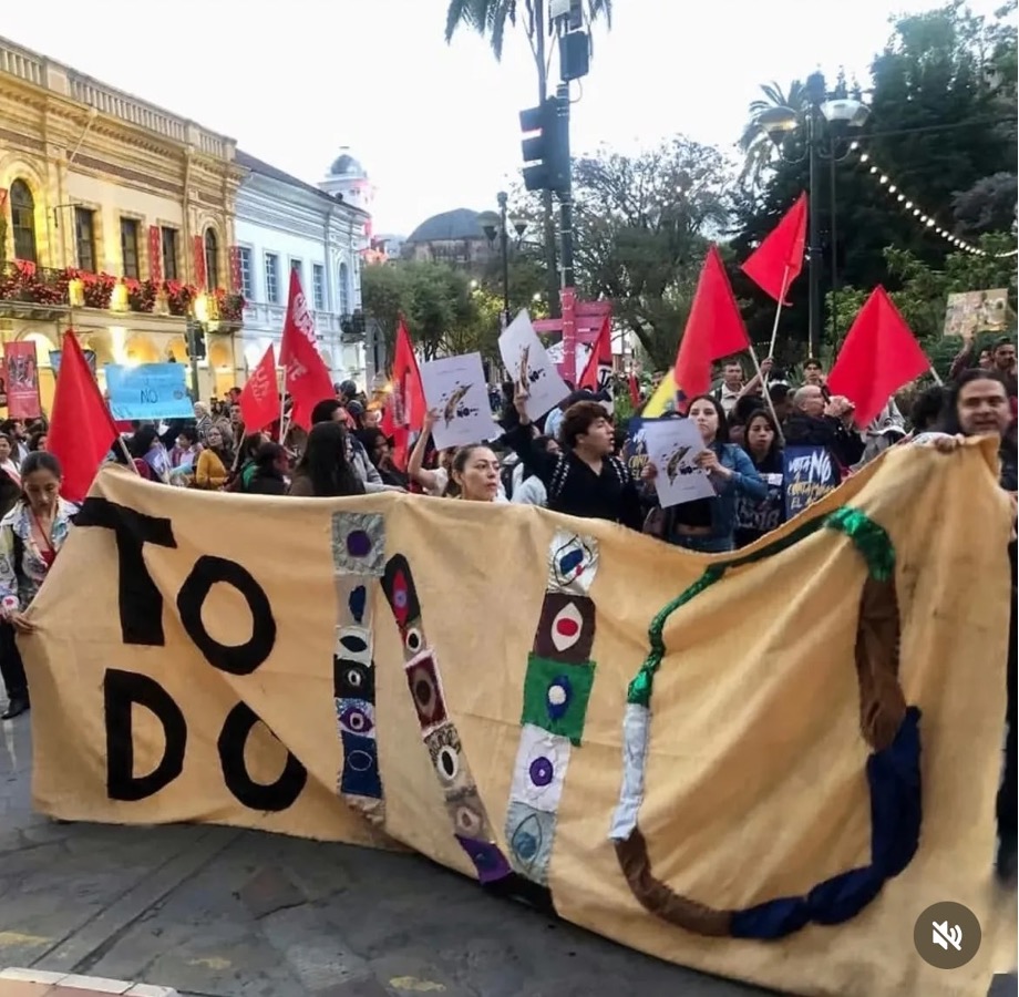 A photo of an outdoor street protest with a large crowd. The people in the foreground hold a large banner made of cloth, but it's hard to read. The people behind them carry signs and red flags.