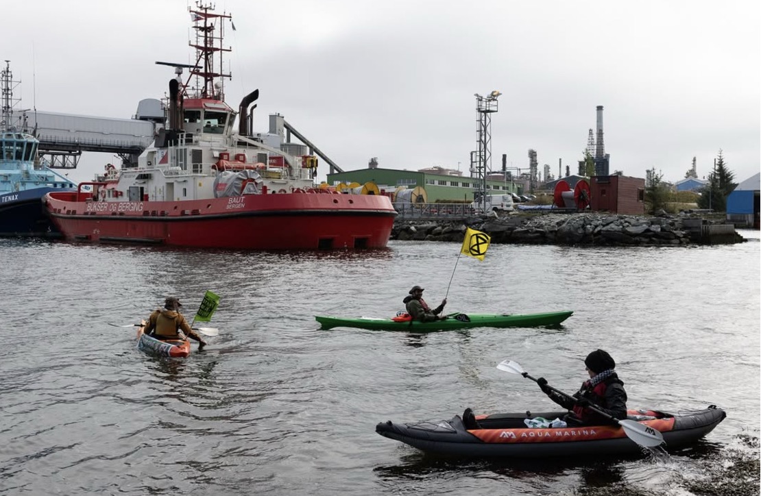 A photograph of what looks like a working harbour under a grey sky. The water is also grey. A large, red-hulled boat is visible in the background, as is part of a blue boat of similar size. In the foreground paddle three humans in kayaks, two of them holding small flags featuring the XR logo.